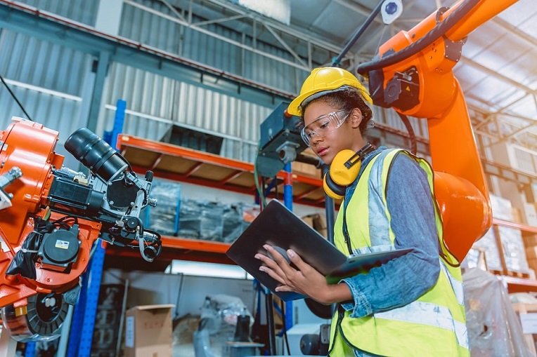 A female engineer in safety gear inspecting machinery with a digital tablet in an industrial setting.