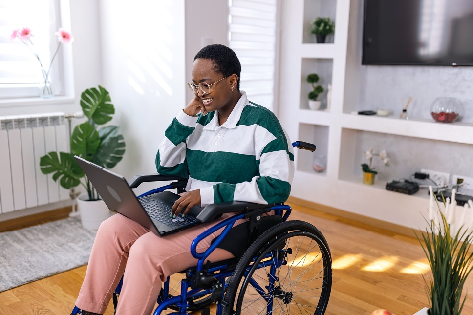 A person in a wheelchair smiling and using a laptop in a cozy room with plants and decor.