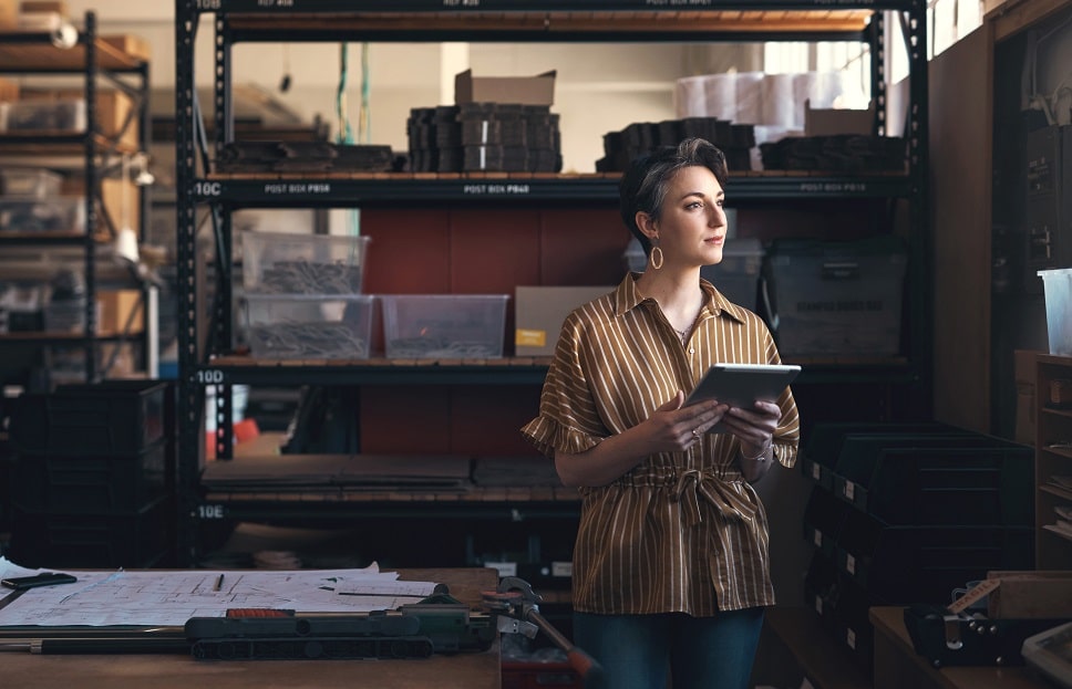A professional woman in a warehouse holding a tablet, possibly managing inventory or production processes.
